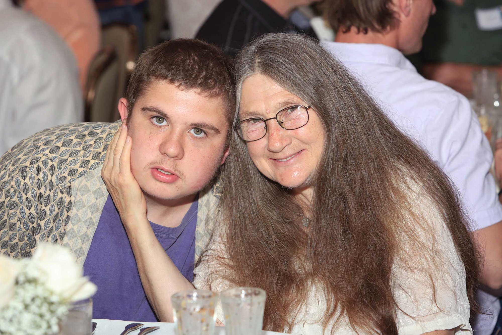 Planned Giving. A man with a disability and a light skin tone sits next to an older woman with long, straight hair. The woman holds her hand against the man’s cheek while smiling toward the camera.