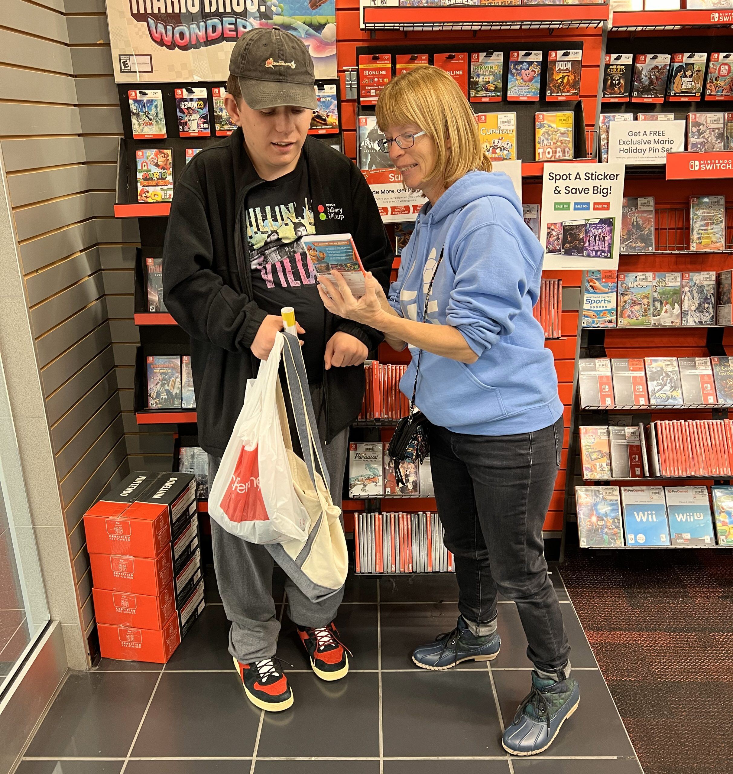 A man with a disability, a light skin tone and short brown hair reads the back of a video game case while holding several plastic bags. next to him is a middle-aged woman with a light skin tone and chin-length blond hair, holding the video game case toward the man.