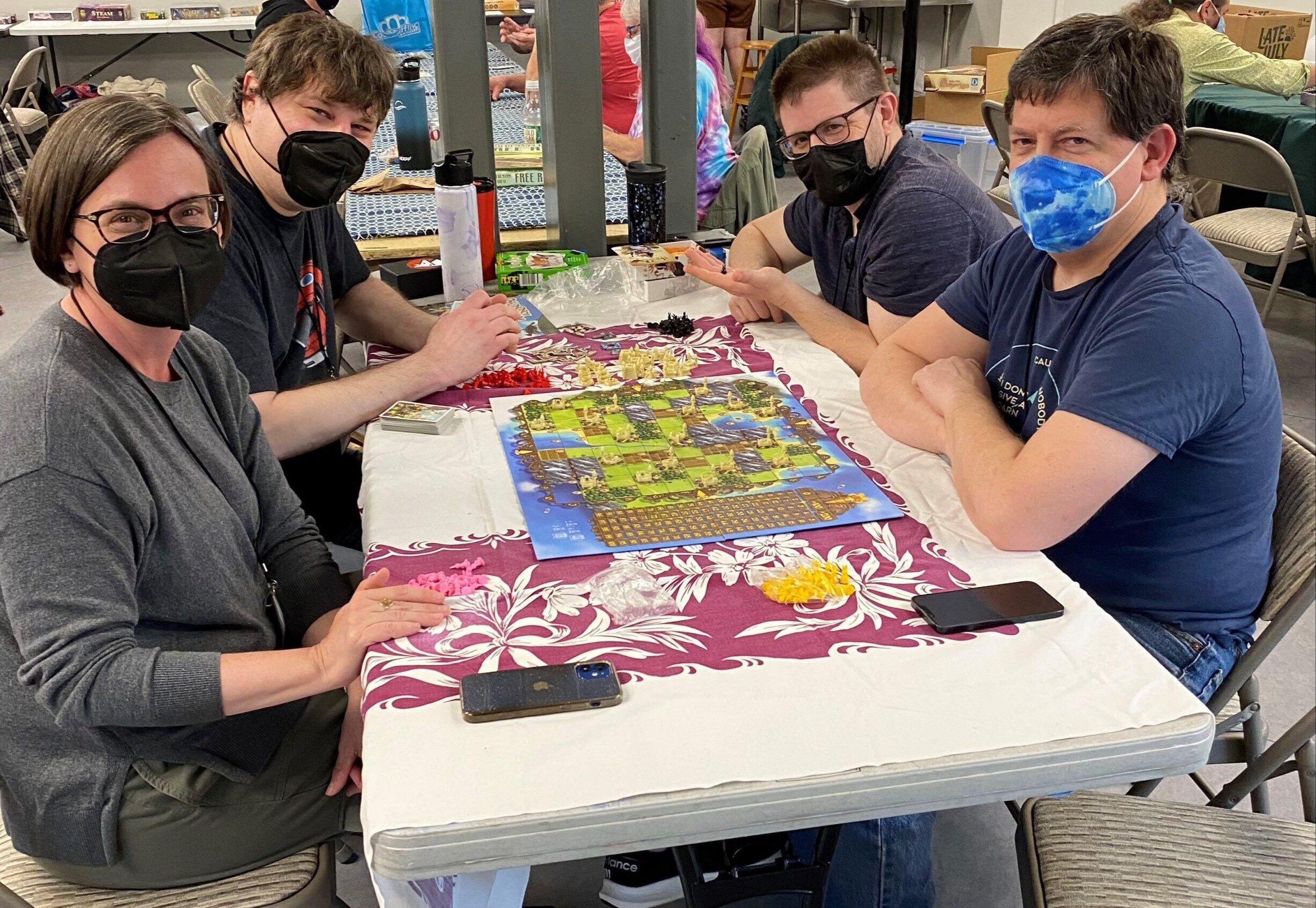 Four people with light skin tones and face masks sit at a table and smile at the camera while playing a board game.