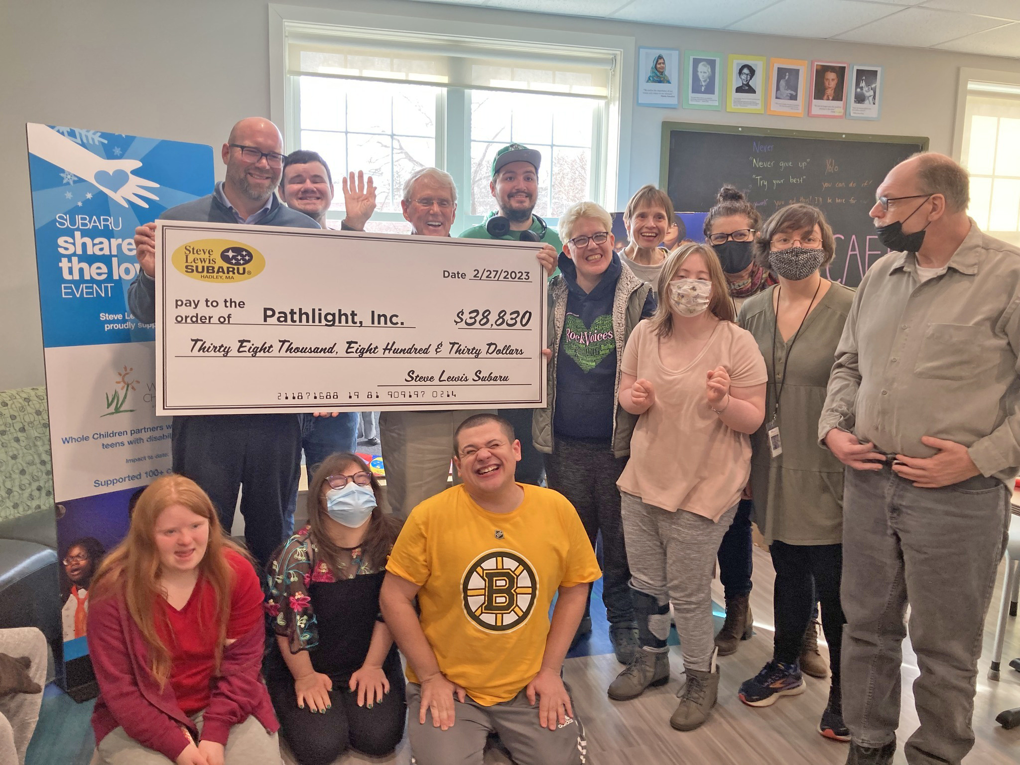 A group of people with light skin tones smile as they hold up a check for $38,830 written out to Pathlight Inc. Some people stand next to the check, others sit in front of it.