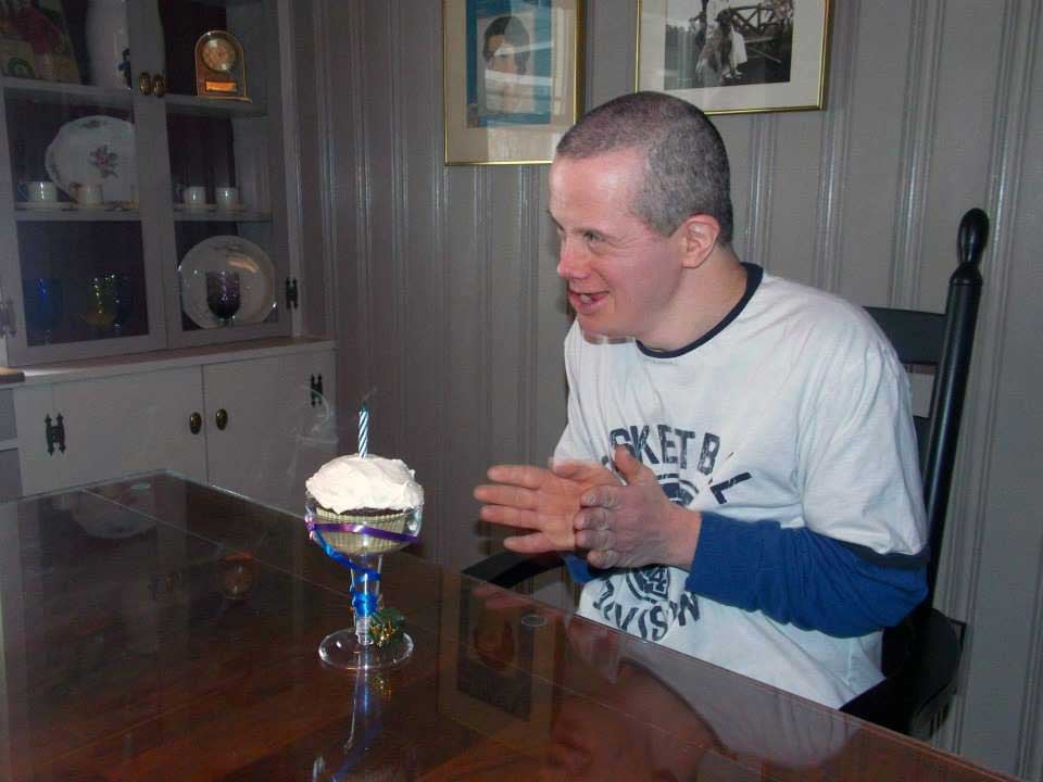 Give happiness. Darren Harington, a man with a light skin tone and Down Syndrome, smiles and claps while sitting in front of a birthday cupcake with a blown-out candle. 