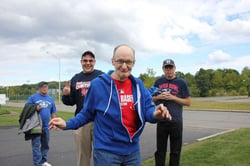 Four men with light skin tones pose for a photo in a parking lot. The man with a disability in front is smiling and wearing a red baseball T-shirt and blue sweater. Behind him on the left is a man holding up the peace sign with his hands while smiling; next to him is a man holding a blue jacket. In the back on the left is a man wearing a blue shirt while carrying a grey coat.