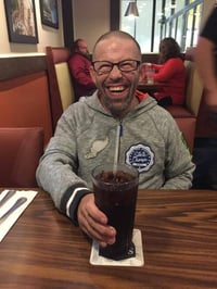 Gerry, a man with a disability and light skin tone, smiles as he looks at the camera while holding a glass of soda.