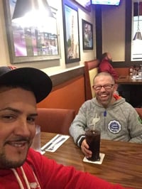 Edwin, a middle-aged man with a medium skin tone, smiles at the camera while he takes a self-portrait with Gerry, a man with a disability and light skin tone, who smiles while holding a glass of soda.