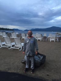 Gerry, a man with a disability and light skin tone, stands on a sandy beach with rows of beach recliners. A lake and mountains are in the distant background.