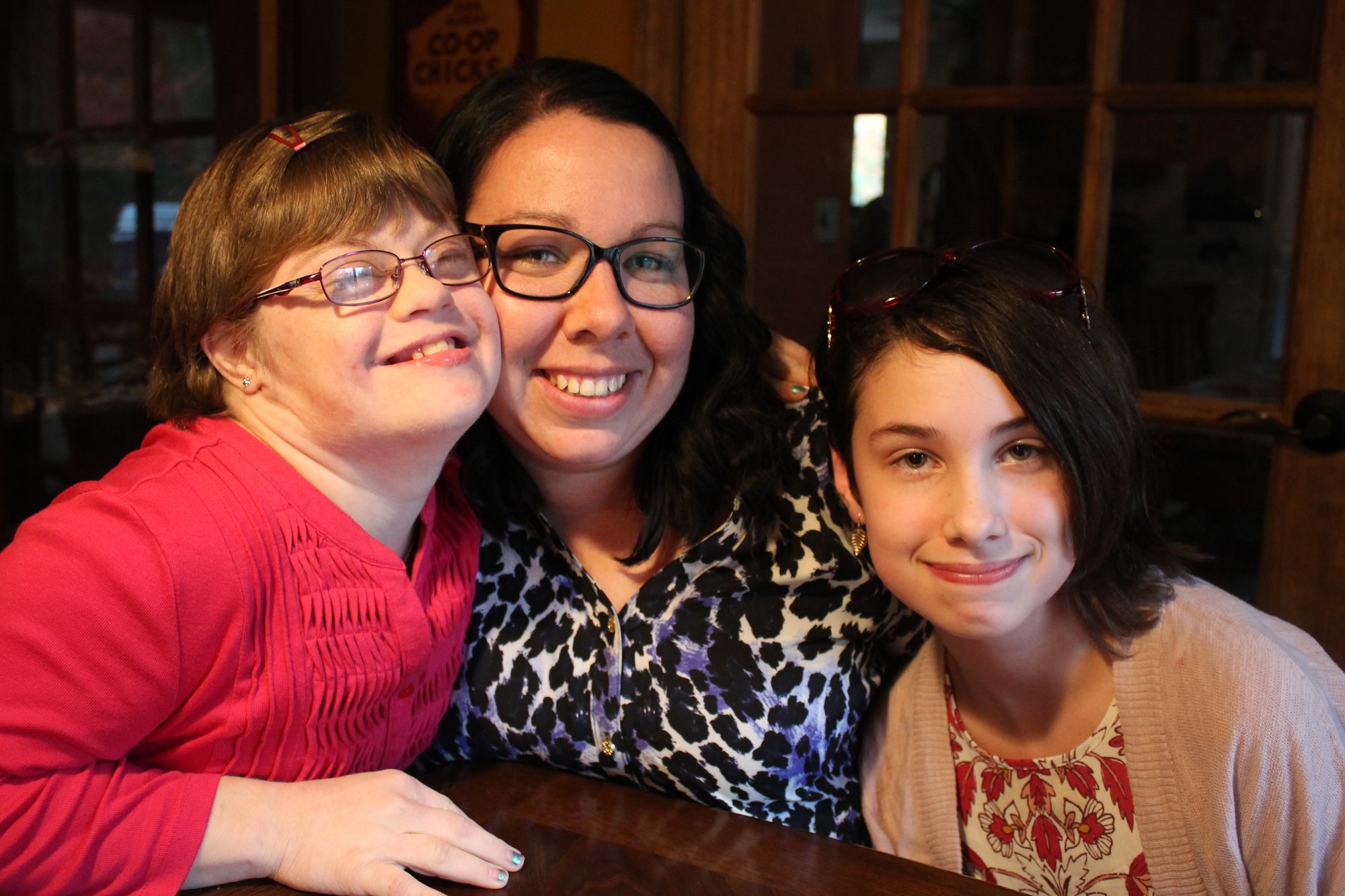 Champion Dignity. Three people with light skin tones sit at a table, smiling at the camera. On the left, a younger girl with Down Syndrome presses her face against another adult woman in the middle. Next to her, on the right, is a smiling teenager.