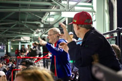 Two older men with light skins in a profile angle wave their hands in the upper stands of Fenway Park during a Boston Red Sox game.