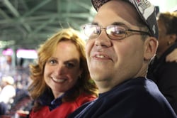 A man and a woman with light skin tones are smiling at the camera while sitting in the Red Sox Stadium. 