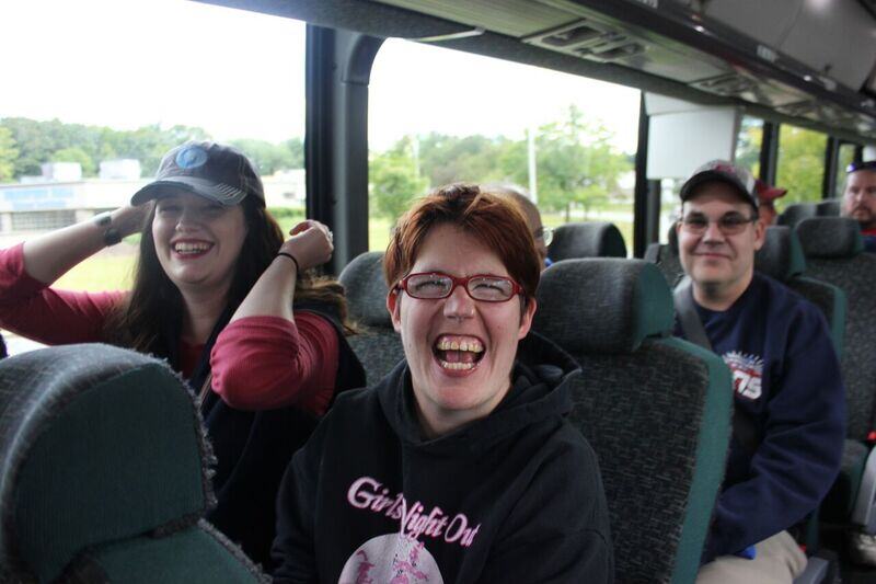 Three people with light skin tones sit on a commercial bus to Fenway Park. The woman in front is laughing with a big smile. Sitting next to her is a smiling woman adjusting her baseball cap. Behind the two women is an older man who is smiling gently.