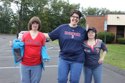 Three women with disabilities and light skin tones pose together for a photograph. The woman on the left is wearing a red Red Sox top and taking off a blue sweater. The woman in the middle is wearing a navy blue BoSox top while smiling wide with her hands around the shoulders of the other two women. The woman on the right is smiling and holding her hand on her hip while wearing a Red Sox top and baseball cap.