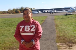 Jerrilis, a woman with a disability and a medium skin tone, stands in a grass patch of a small airport. In the background are several white, light-weight planes.