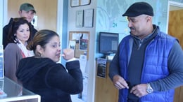 Jerrilis, a woman with a disability and a medium skin tone, looks at an older man wearing a blue vest. Behind Jerrilis is another young woman looking at both of them.