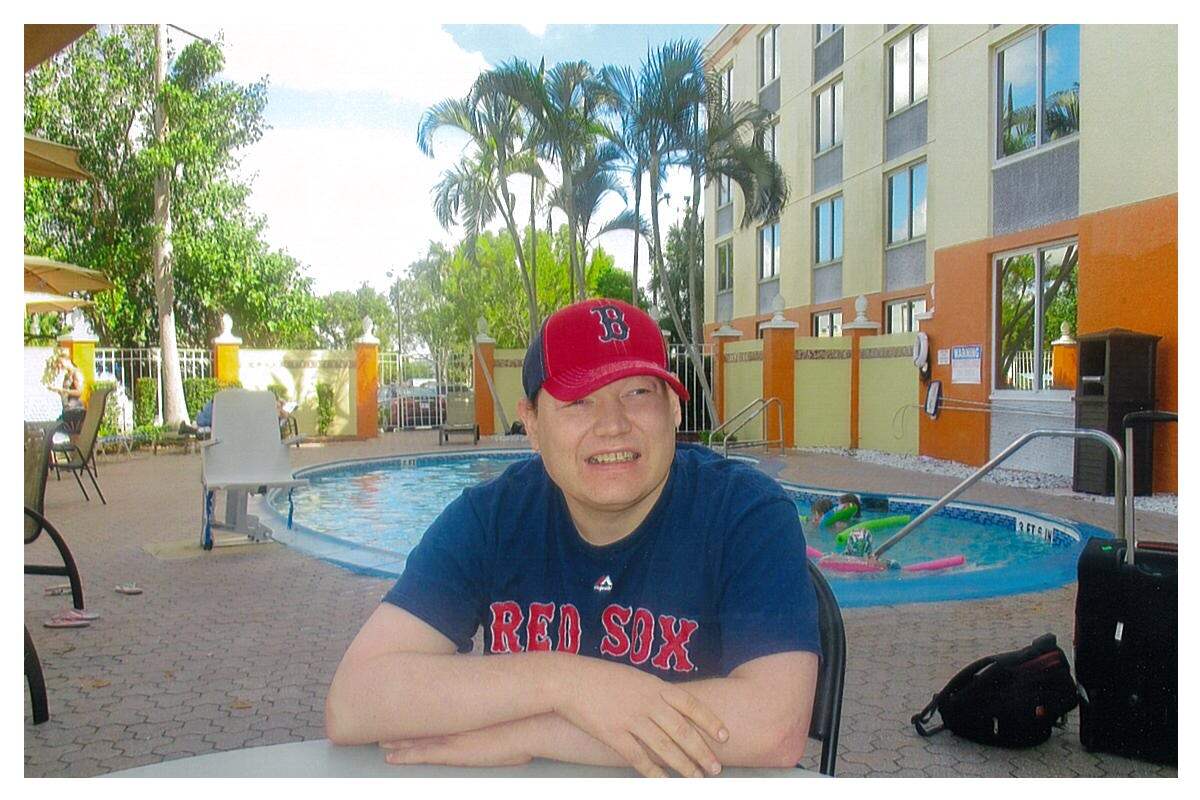 Michael, a man with a disability and a light skin tone, sits at a table in front of a hotel pool. He is smiling and wearing Boston Red Sox merchandise.