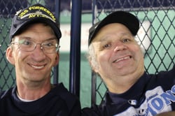 Portrait of two men with disabilities and light skin tones smiling at the camera in front of a fence. 