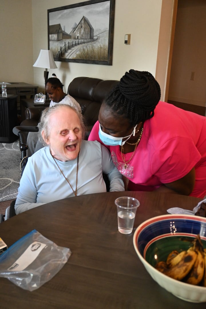 Residential Supports. An older woman with a light skin tone in a wheelchair laughs while sitting at a table. A nurse with a dark skin tone in bright pink scrubs and a face mask leans over to speak with her. 