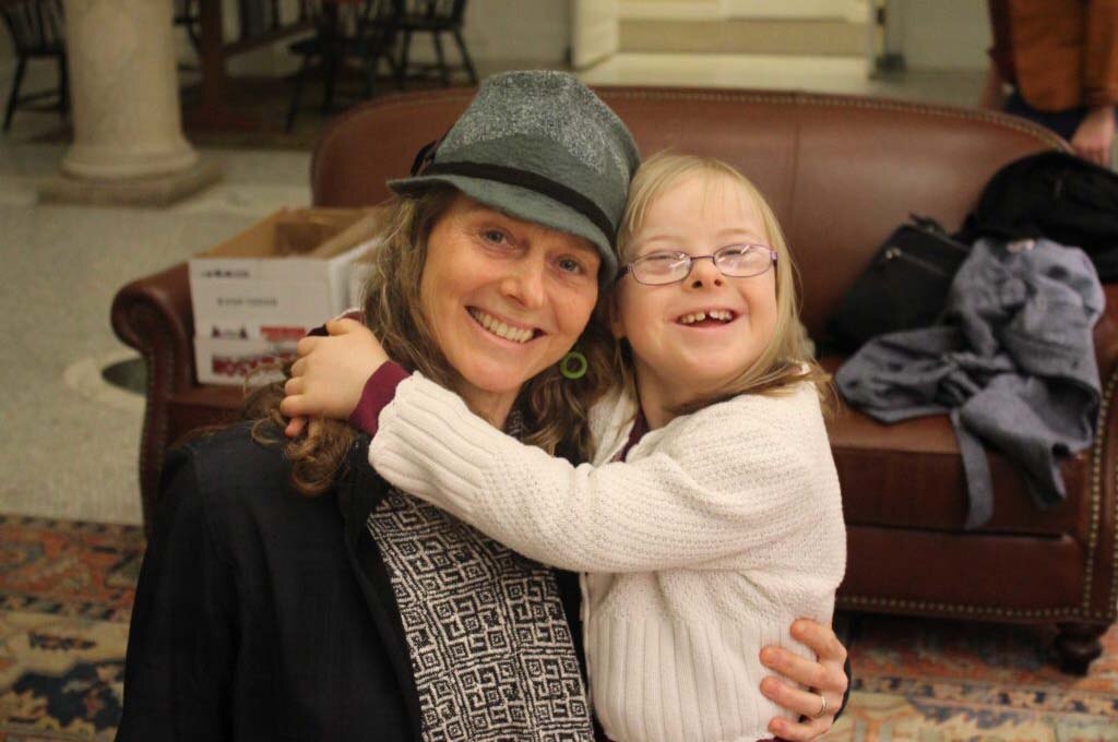 Down Syndrome Resource Group of Western Massachusetts. A girl with Down Syndrome and a light skin tone smiles as she hugs a woman with a light skin tone, wearing a grey trilby. The woman holds the girl with one arm and smiles at the camera.