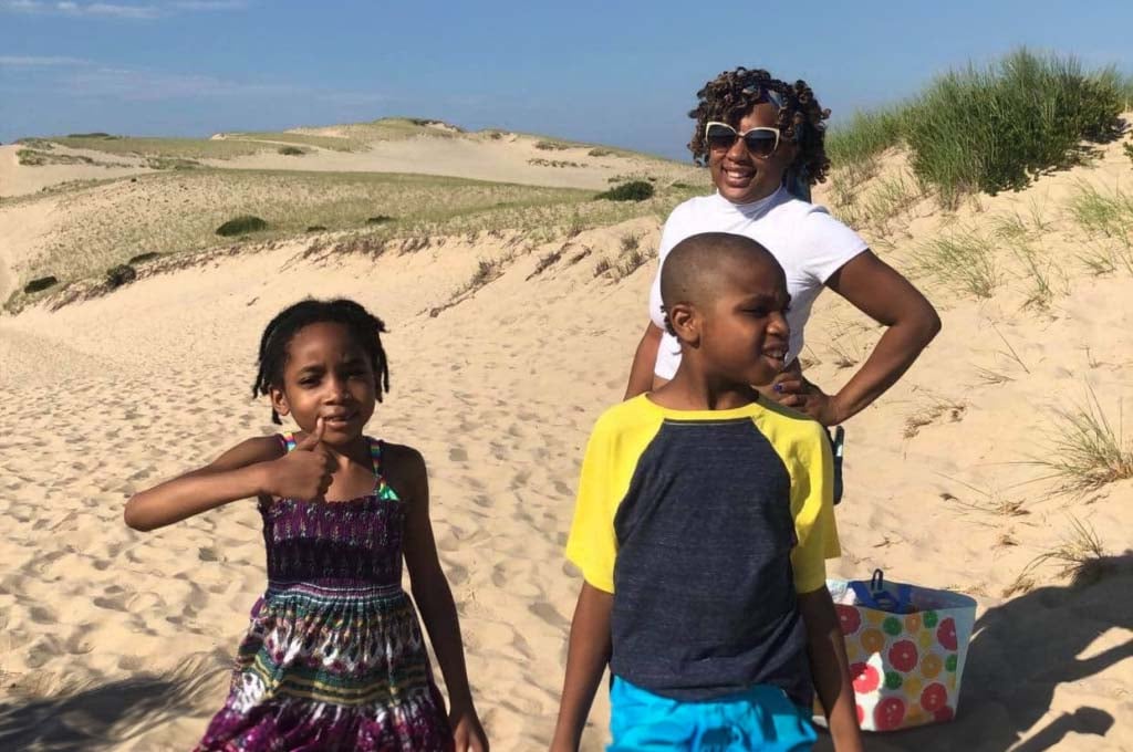 Family Empowerment. In front of grassy dunes, a woman with a dark skin tone is wearing glasses with her hand on her hip and smiles. On the left in the foreground, a girl with a dark skin tone gives a thumbs up while looking at the camera. On the right, a boy with a dark skin tone squints as he looks off to the side.