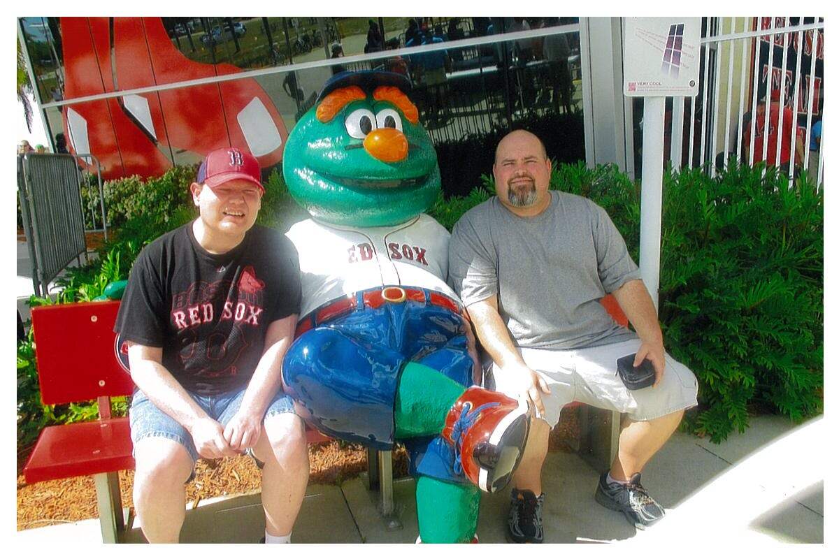 Two men with light skin tones sit beside the Boston Red Sox mascot statue, Wally the Green Monster. The man with a disability seated on the left is wearing Boston Red Sox fan gear while squinting and smiling. The man sitting on the right is smiling.