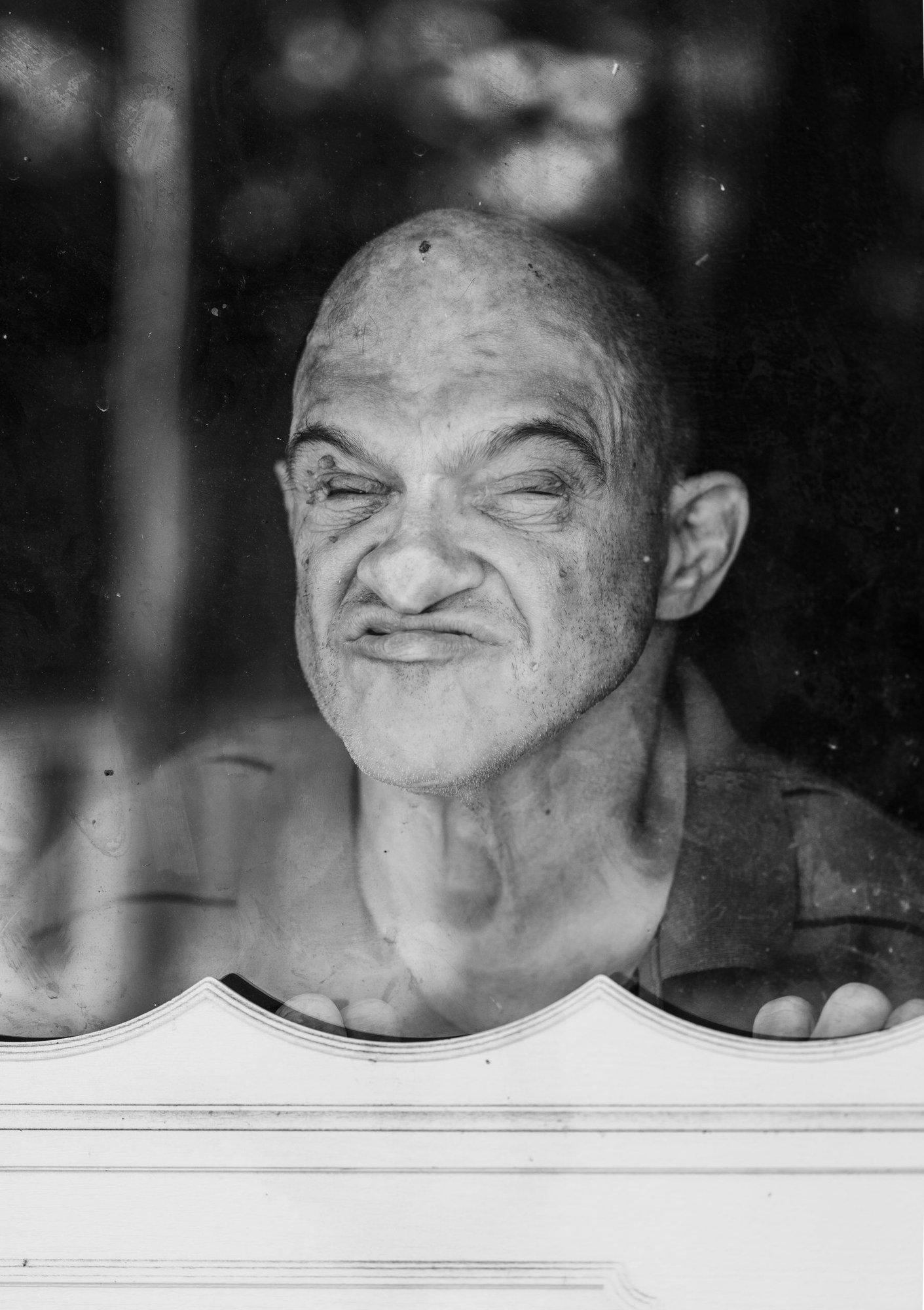 A black and white portrait of a disabled man with a light skin tone pressing his nose against a window while squinting at the camera.