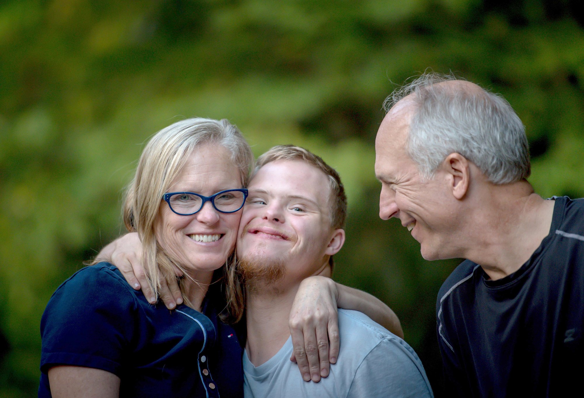 Autism Connections. A middle-aged woman with a light skin tone and a man with Down Syndrome and light skin tone smile as they hang their arms around each other's shoulders. On the right, an older man with a light skin tone smiles at both.