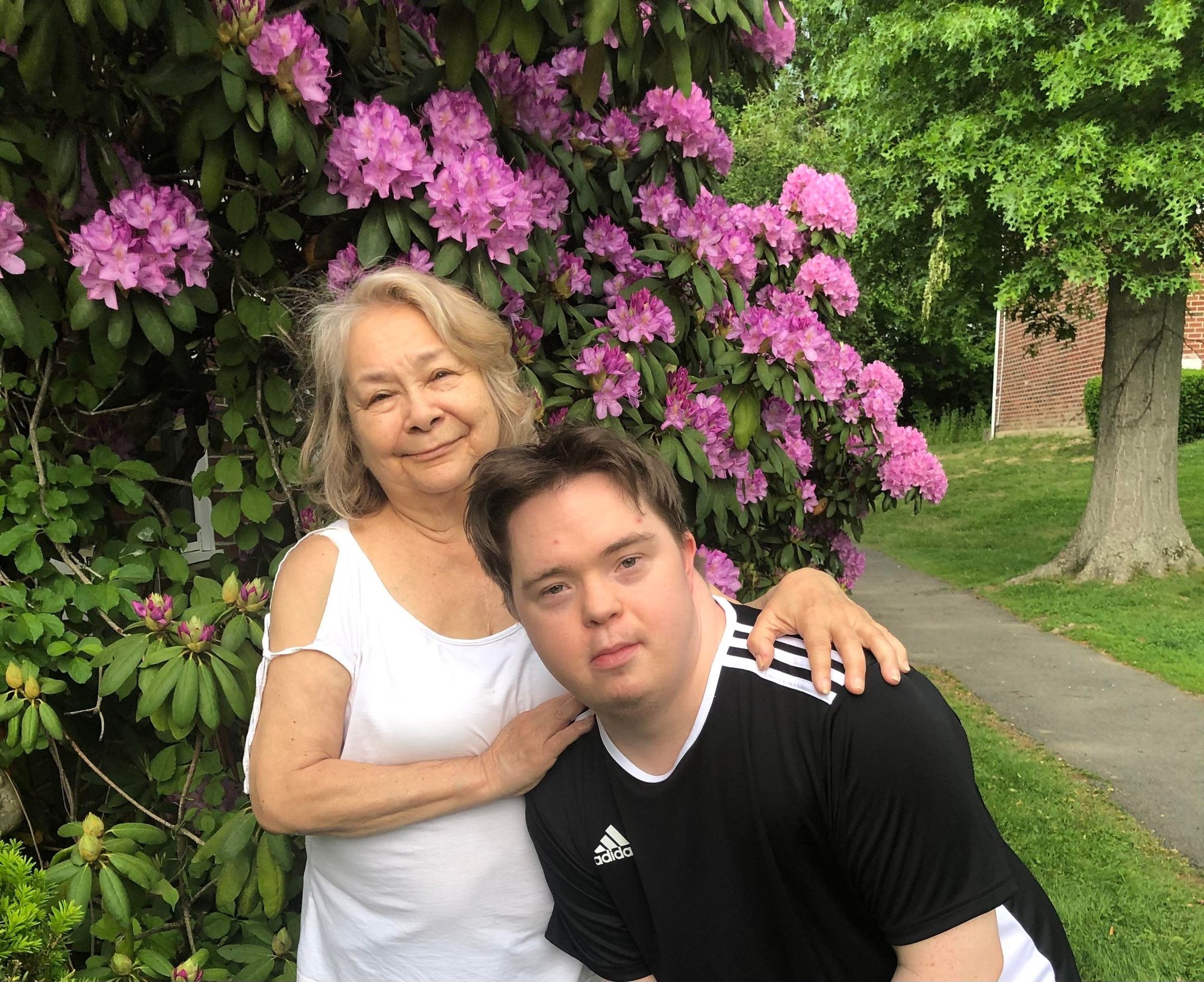 Adult Family/Foster Care. A man with Down Syndrome and a light skin tone is leaning over in front of an older woman with a light skin tone and white hair. She keeps her hands on his shoulder while smiling. They are both standing in front of a flowering bush.