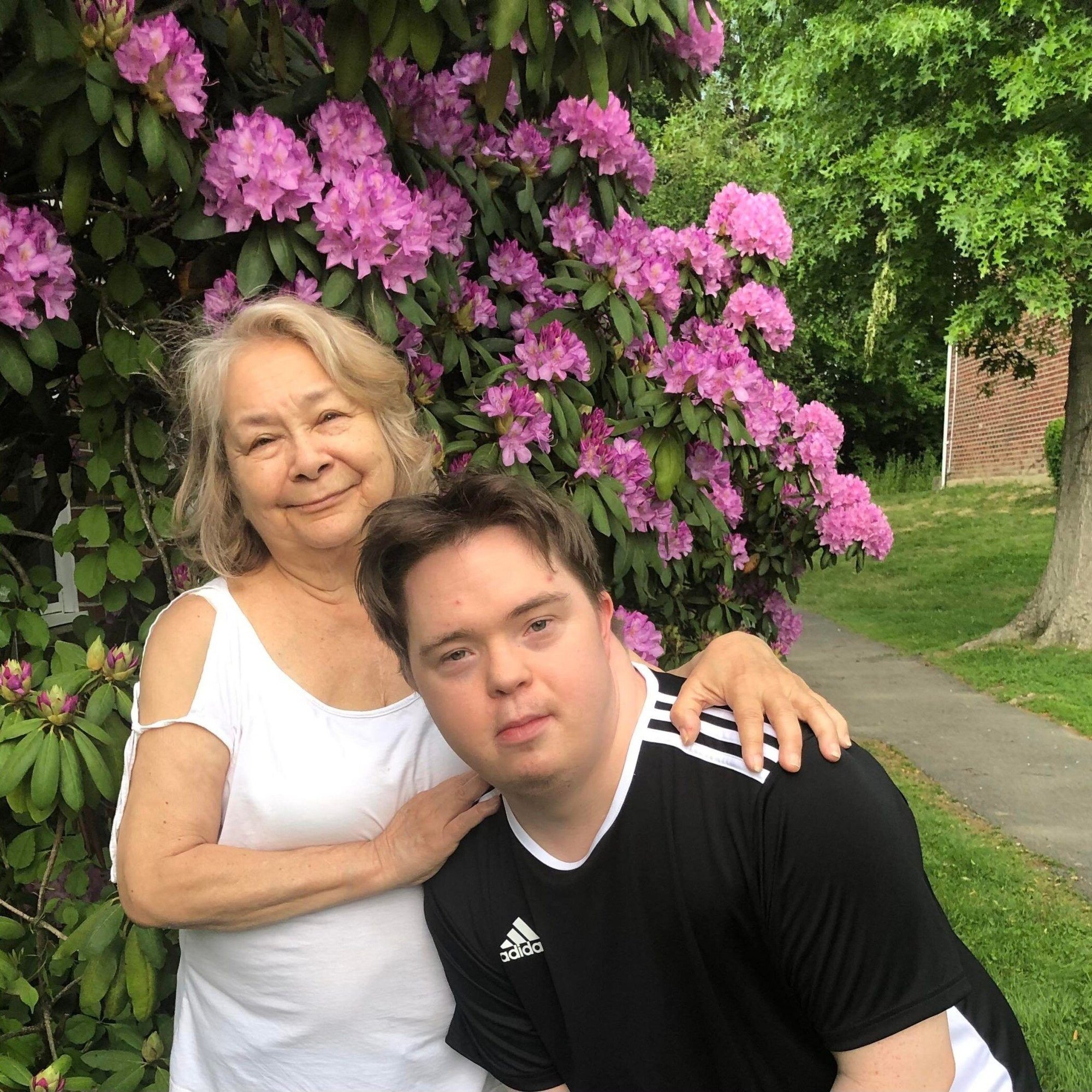 Adult Family/Foster Care. A man with Down Syndrome and a light skin tone is leaning over in front of an older woman with a light skin tone and white hair. She keeps her hands on his shoulder while smiling. They are both standing in front of a flowering bush.