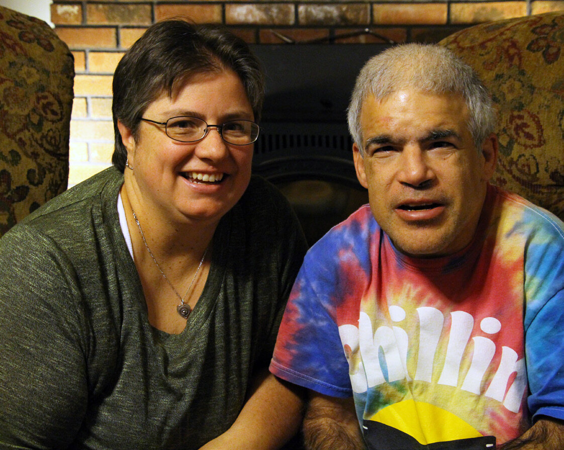 Shared Living. A woman with a light skin tone and short black hair smiles at the camera on the left side. On the right, an older man with a medium skin tone is wearing a tie-dye shirt with the text, chilling, and sits beside the woman while looking at the camera.