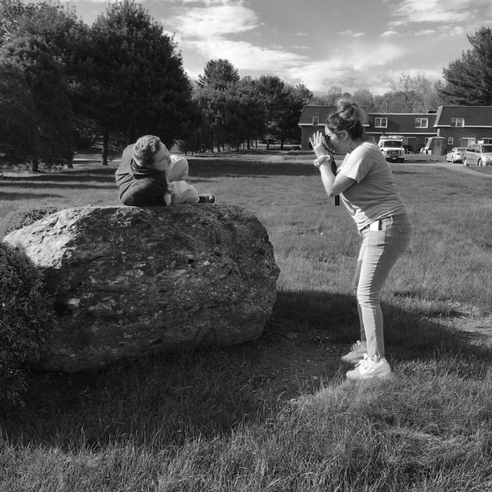 In black and white, a photographer with a light skin tone takes a photo of a man with a disability and light skin tone lying on a boulder near the previous Inclusive Community Center in Hadley.