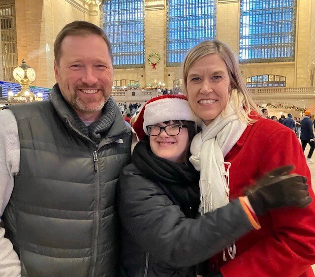 Three people with light skin tones smile as they pose for a photo. The woman with a disability in the center is wearing a Santa Claus hat while hugging a middle-aged woman with a red coat. The middle-aged man on the left is wearing a vest.