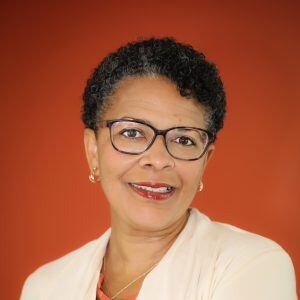 A portrait of Pathlight Board President Denise Cogman, a middle-aged woman with a middle-dark skin tone, short black hair, and glasses.