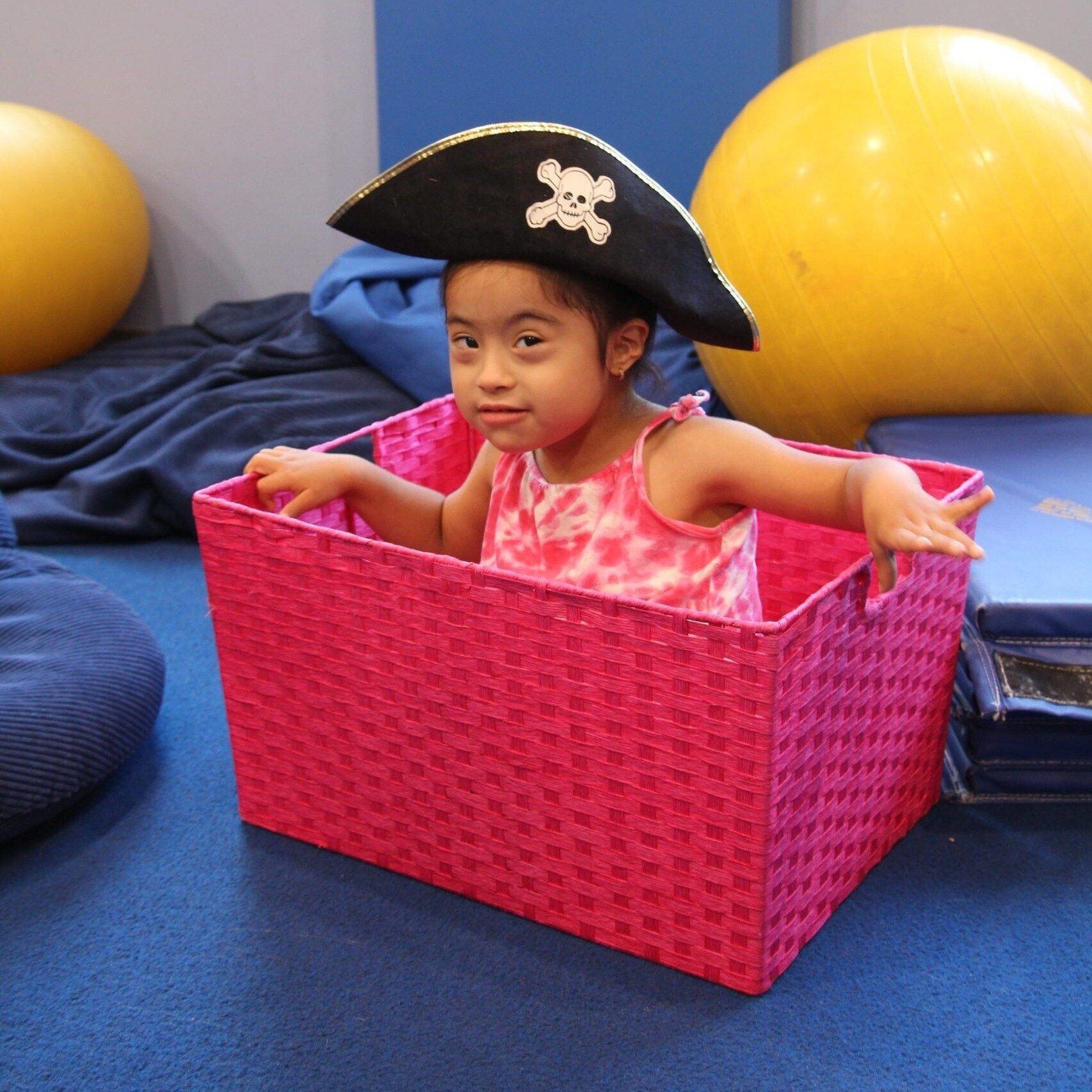 A girl with Down Syndrome and a medium skin tone wears a pirate hat while sitting in a large pink basket.